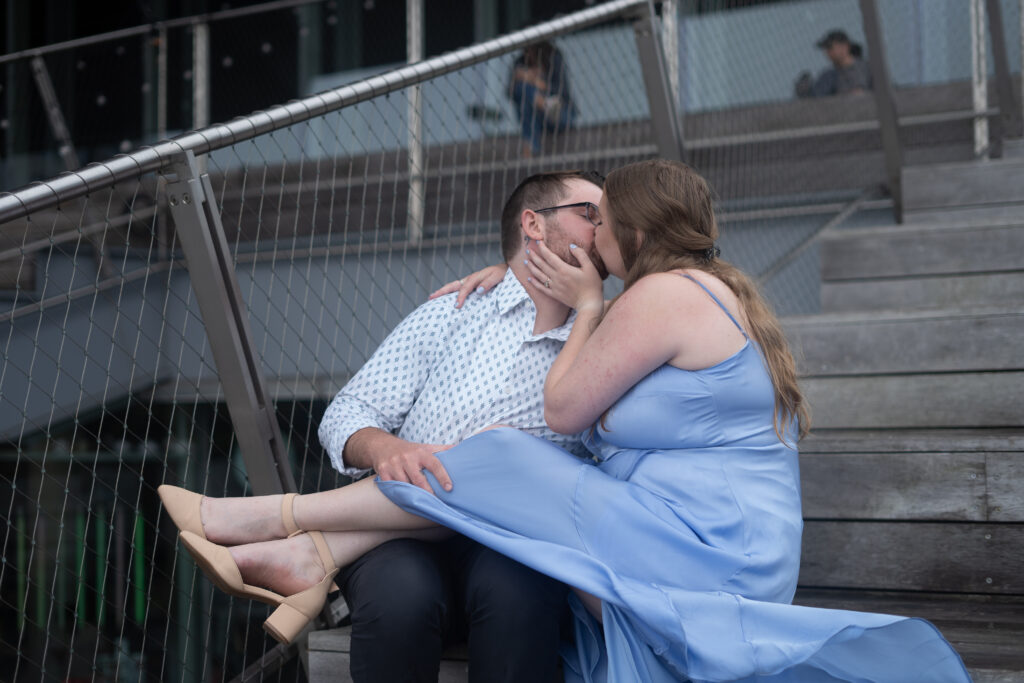 A loving couple embraces in the heart of Boston's Seaport District, surrounded by the city skyline and waterfront. The raw, unedited image captures the genuine joy and connection between them, as they share a moment of laughter and intimacy. The natural light highlights the details of their expressions, and the background features iconic landmarks, creating a timeless and authentic engagement photo.