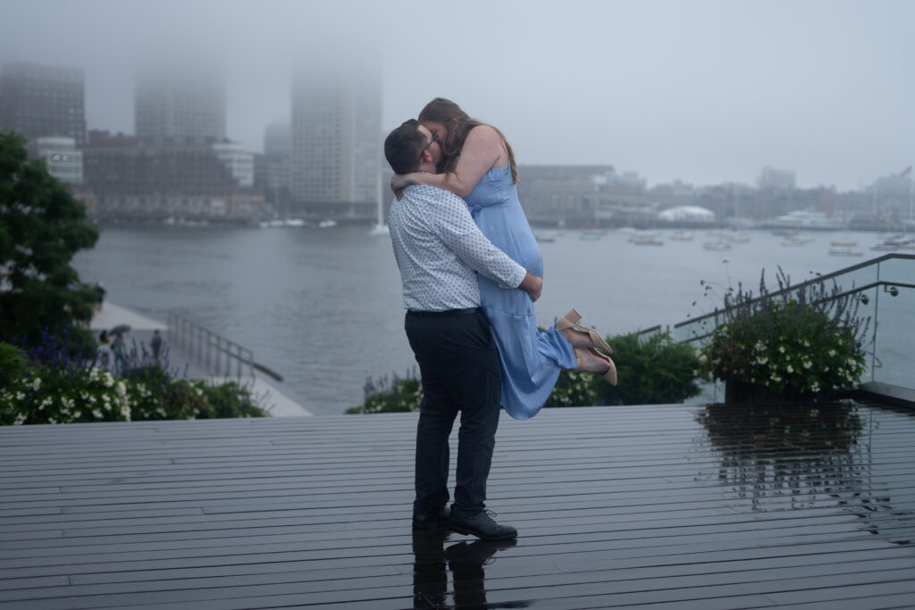 A loving couple embraces in the heart of Boston's Seaport District, surrounded by the city skyline and waterfront. The raw, unedited image captures the genuine joy and connection between them, as they share a moment of laughter and intimacy. The natural light highlights the details of their expressions, and the background features iconic landmarks, creating a timeless and authentic engagement photo.