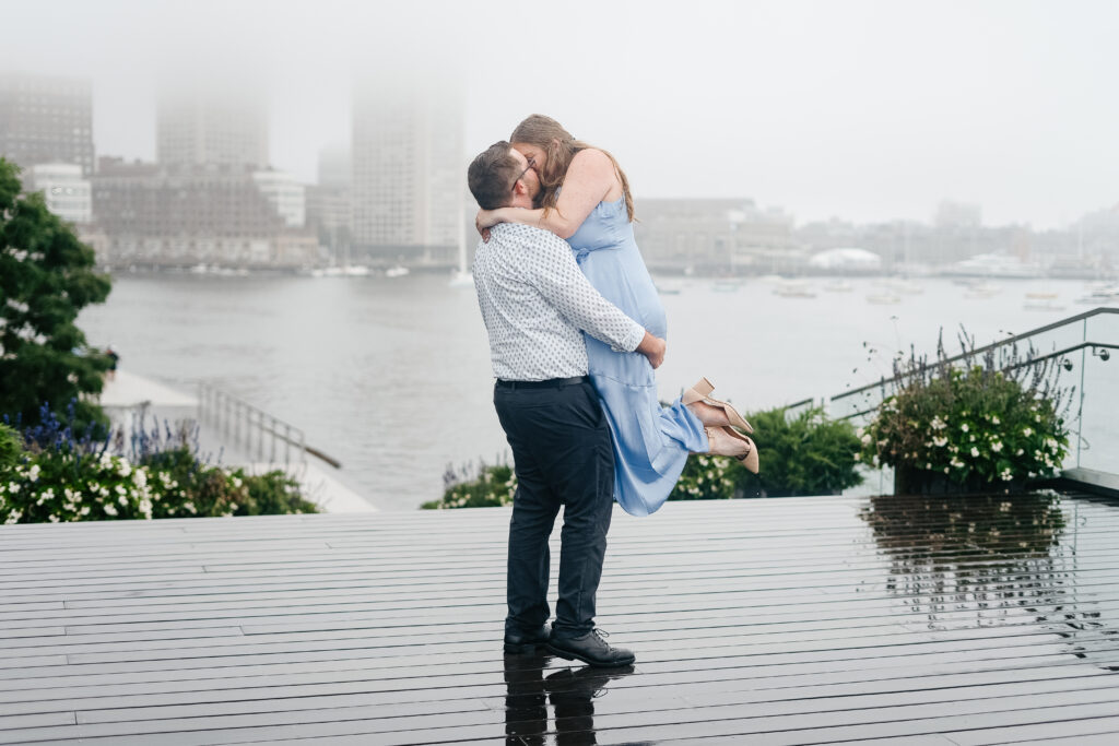 In this enhanced engagement photo, a radiant couple shares a tender embrace against the stunning backdrop of Boston's Seaport District. The carefully edited image features vibrant colors, a seamless blend of natural and enhanced lighting, and a refined background that accentuates the romantic atmosphere. The couple's joy is amplified through subtle skin retouching, bringing out their best features while maintaining the authenticity of the moment. The result is a captivating and polished portrayal of love in the heart of the city.
