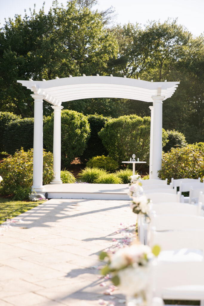 A stunning image of a wedding ceremony at The Villa in Bridgewater, capturing the elegant outdoor setting with manicured gardens and a charming gazebo. The soft, natural light creates a romantic ambiance as the couple exchanges vows, with guests seated on the beautifully arranged lawn. The venue’s classic architecture and picturesque surroundings make it a truly enchanting backdrop for a memorable day.