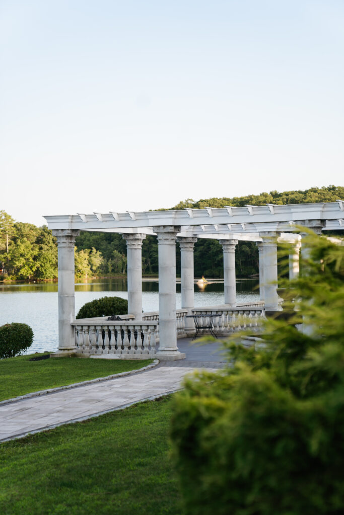 A stunning wide-angle shot of a wedding at Grand View in Mendon, showcasing the expansive, scenic views of the surrounding landscape. The couple stands on a terrace overlooking rolling hills and a tranquil lake, bathed in soft, golden hour light. The venue’s name truly shines through in this breathtaking backdrop, highlighting the beauty and serenity of the location, making it an unforgettable setting for their special day.