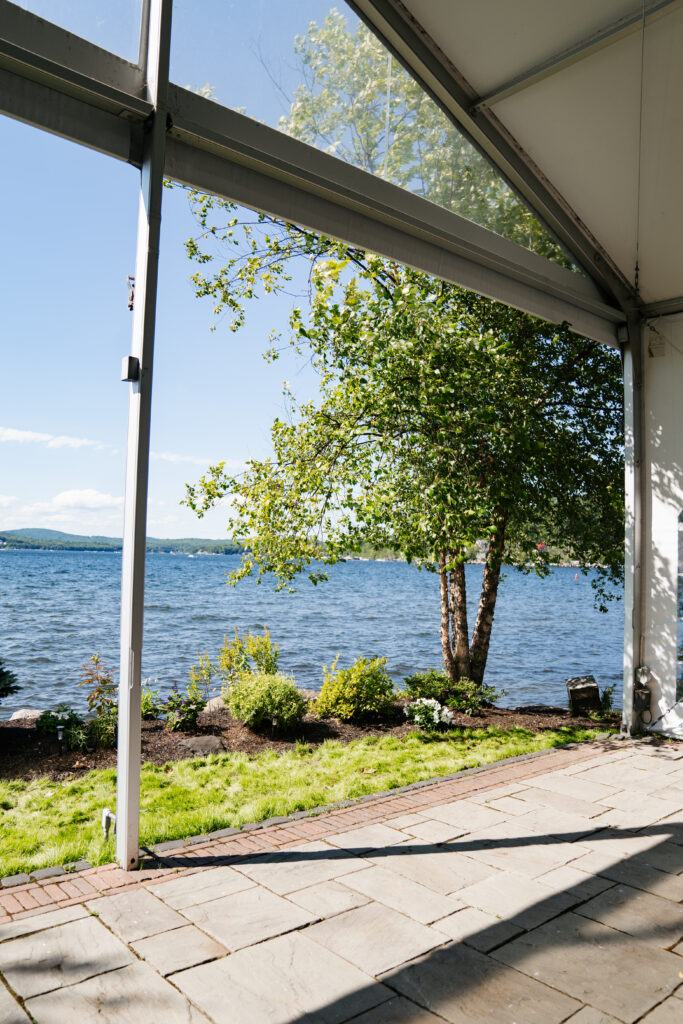 A stunning image of a wedding at The Margate on Lake Winnipesaukee, with the couple standing on the lakeside deck as the sun sets behind them, casting a warm golden glow over the water. The serene, picturesque setting of the lake and surrounding mountains creates a tranquil yet breathtaking backdrop for their special day. The natural beauty of the location, combined with the intimate outdoor setting, captures the essence of a perfect lakeside celebration.