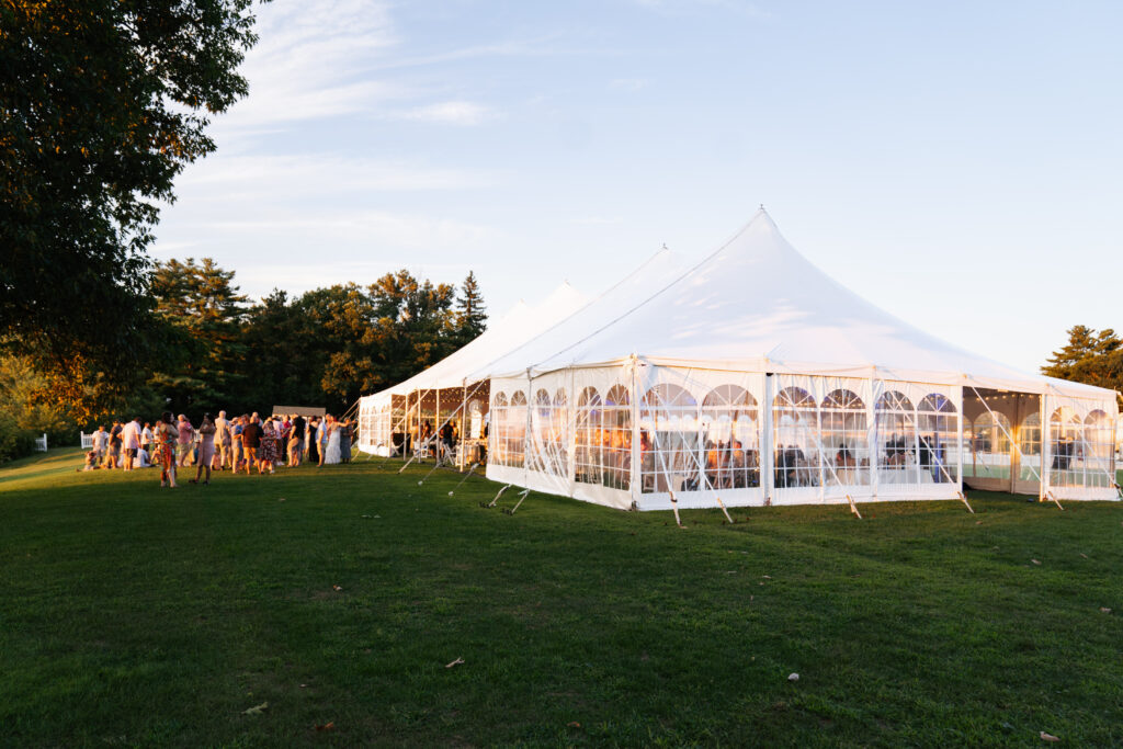 A stunning image of a wedding at The Margate on Lake Winnipesaukee, with the couple standing on the lakeside deck as the sun sets behind them, casting a warm golden glow over the water. The serene, picturesque setting of the lake and surrounding mountains creates a tranquil yet breathtaking backdrop for their special day. The natural beauty of the location, combined with the intimate outdoor setting, captures the essence of a perfect lakeside celebration.